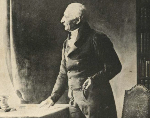 Johann Hermann Kuetemeyer trägt weißes Haar und Anzug, ist um die 70 oder 80 Jahre alt und steht in einer Schwarz-Weiß-Aufnahme vor einem Schreibtisch.