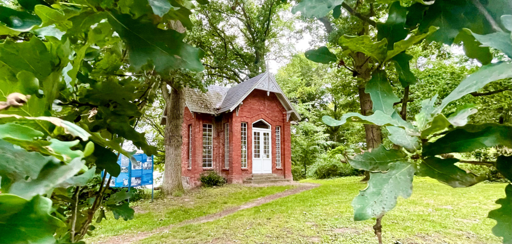 Ein kleines Backsteinhaus mit hohen Fenstern und einer weißen Eingangstür. Es steht im Park, umgeben von viel Grün. Neben dem Häuschen steht eine Schautafel.