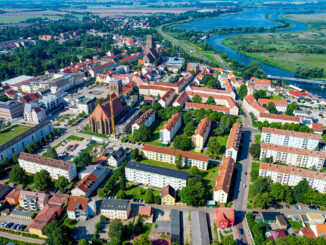 Eine Luftaufnahme von Anklam. Darauf sind viele Häuser, zwei Kirchen, Bäume und ein Fluss. Über den Fluss führt eine große Brücke.