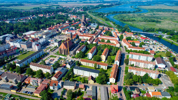 Eine Luftaufnahme von Anklam. Darauf sind viele Häuser, zwei Kirchen, Bäume und ein Fluss. Über den Fluss führt eine große Brücke.