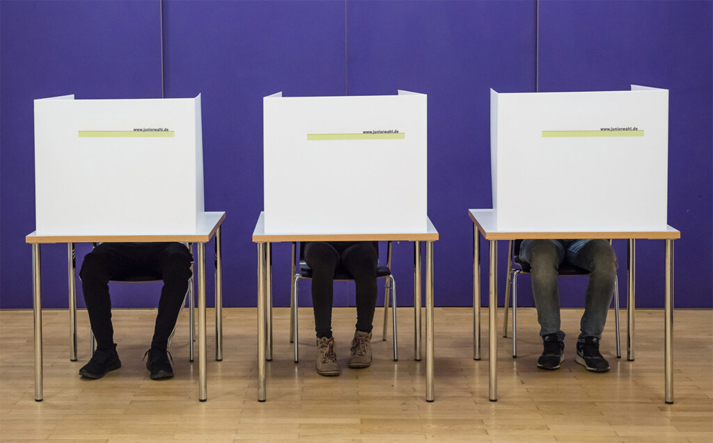 Drei Tische. Auf den Tischen stehen Wahlurnen. Hinter den Wahlurnen sitzen Kinder. Von ihnen sieht man nur die Beine.