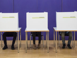 Drei Tische. Auf den Tischen stehen Wahlurnen. Hinter den Wahlurnen sitzen Kinder. Von ihnen sieht man nur die Beine.