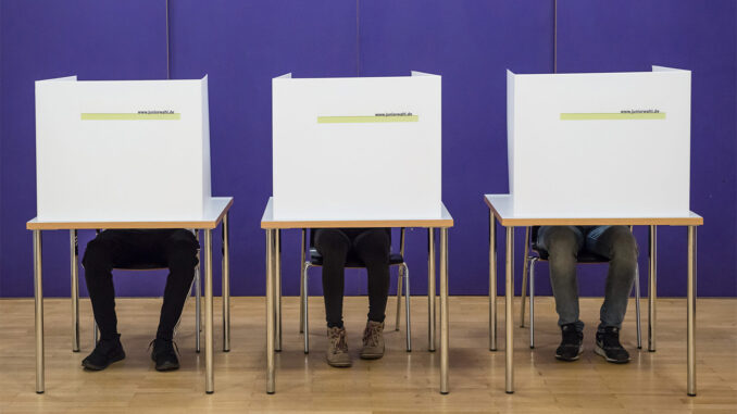 Drei Tische. Auf den Tischen stehen Wahlurnen. Hinter den Wahlurnen sitzen Kinder. Von ihnen sieht man nur die Beine.